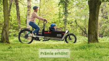 Eine Frau mit Kind auf einem Lastenrad, die durch einen Park fährt. Hinweis Prämie erhöht.